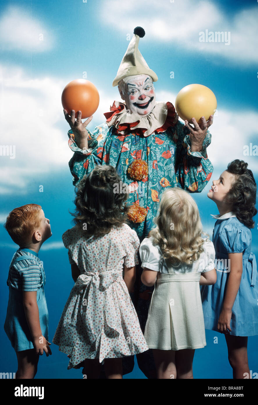 1940s 1950s GROUP OF CHILDREN WATCHING A CLOWN HANDLE ORANGE AND YELLOW BALLS Stock Photo