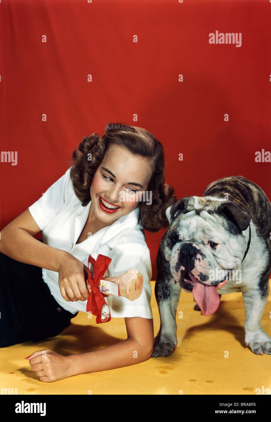 1940s 1950s YOUNG WOMAN GIVING BULLDOG A PRESENT OF A BONE WRAPPED IN RED BOW Stock Photo