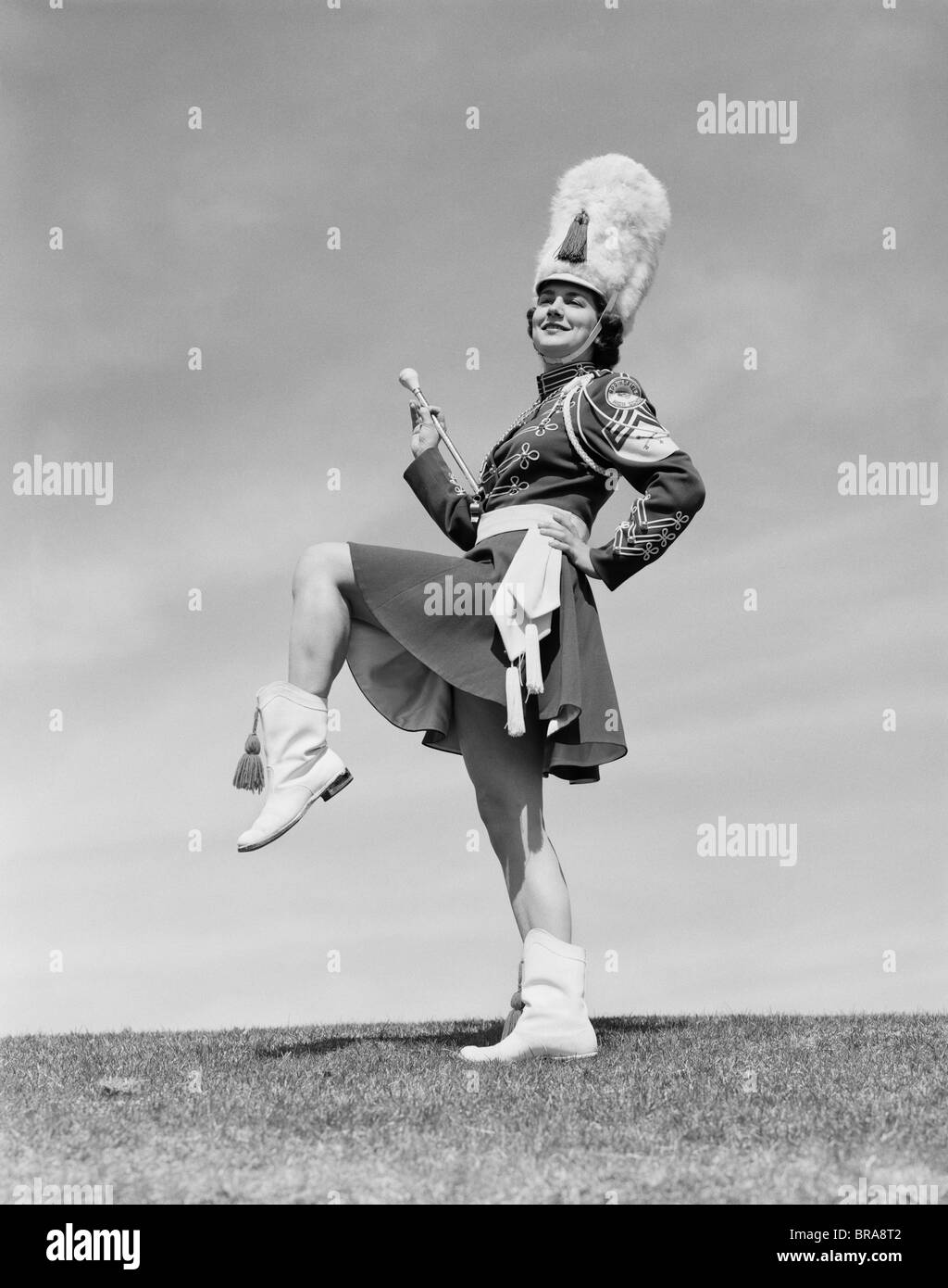 1950s SMILING MAJORETTE POSING OUTDOORS WEARING UNIFORM WITH SHORT SKIRT BOOTS TALL FURRY HAT HOLDING A BATON Stock Photo