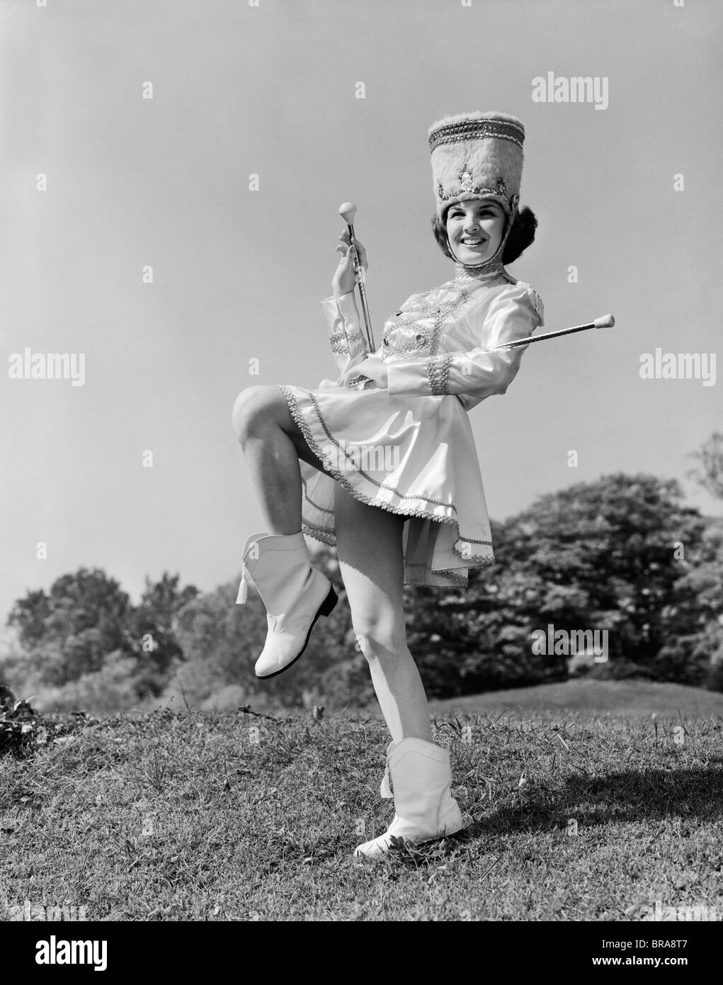 1960s SMILING MAJORETTE IN UNIFORM WITH SHORT SKIRT BOOTS TALL HAT AND TWO BATONS POSING OUTDOORS Stock Photo