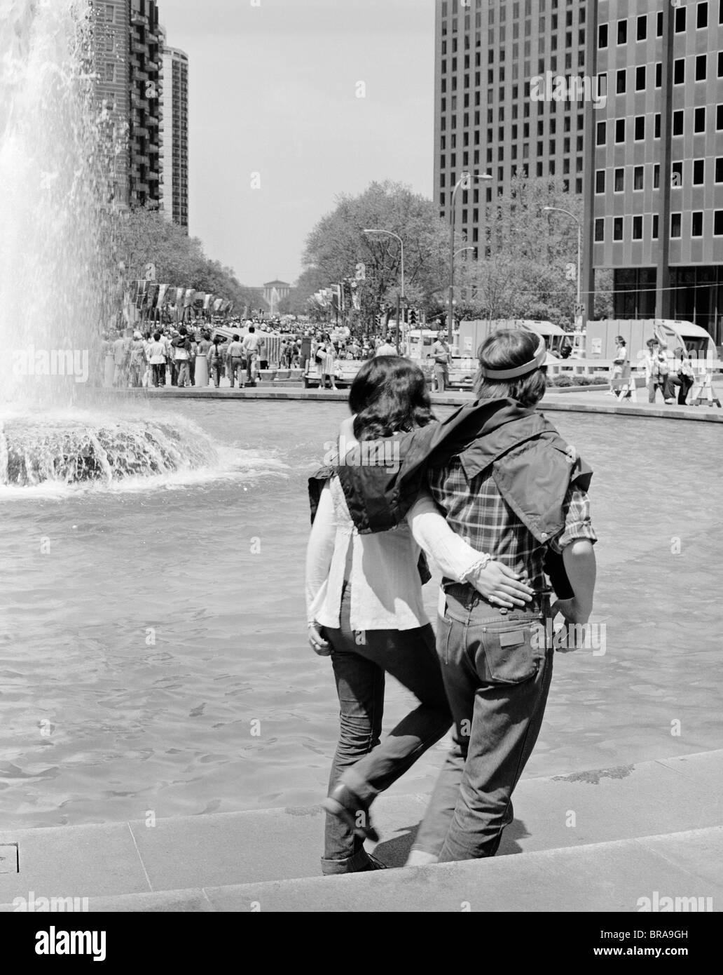 1970s ROMANTIC YOUNG HIPPIE COUPLE WALKING ARM IN ARM BESIDE FOUNTAIN Stock Photo