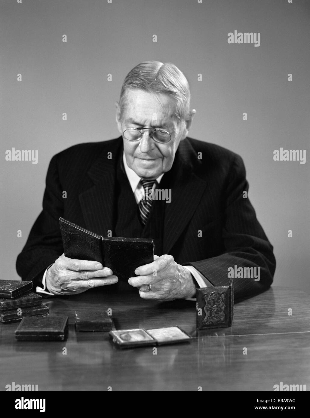 1940s ELDERLY MAN WEARING GLASSES SITTING AT TABLE LOOKING AT DAGUERREOTYPE PHOTOGRAPHS Stock Photo