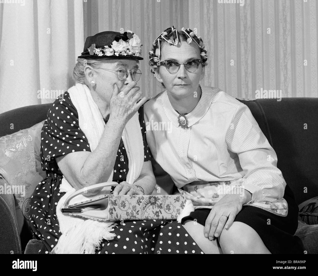 1960s OLDER WOMEN ON COUCH SHARING SECRET ONE IN FLOWERED HAT ONE IN HAIR CURLERS Stock Photo