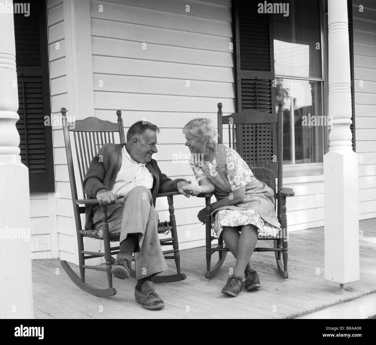1970s ELDERLY COUPLE IN ROCKING CHAIRS ON PORCH HOLDING HANDS Stock Photo