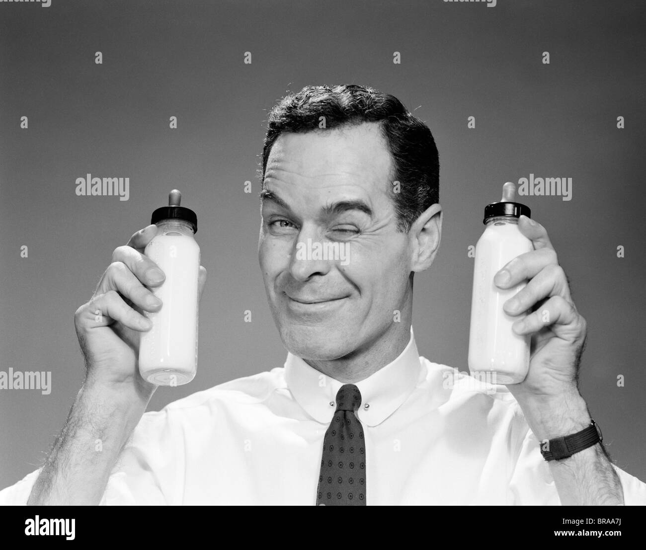 1960s MAN IN SHIRT AND TIE WINKING AT CAMERA HOLDING TWO BABY MILK BOTTLES Stock Photo