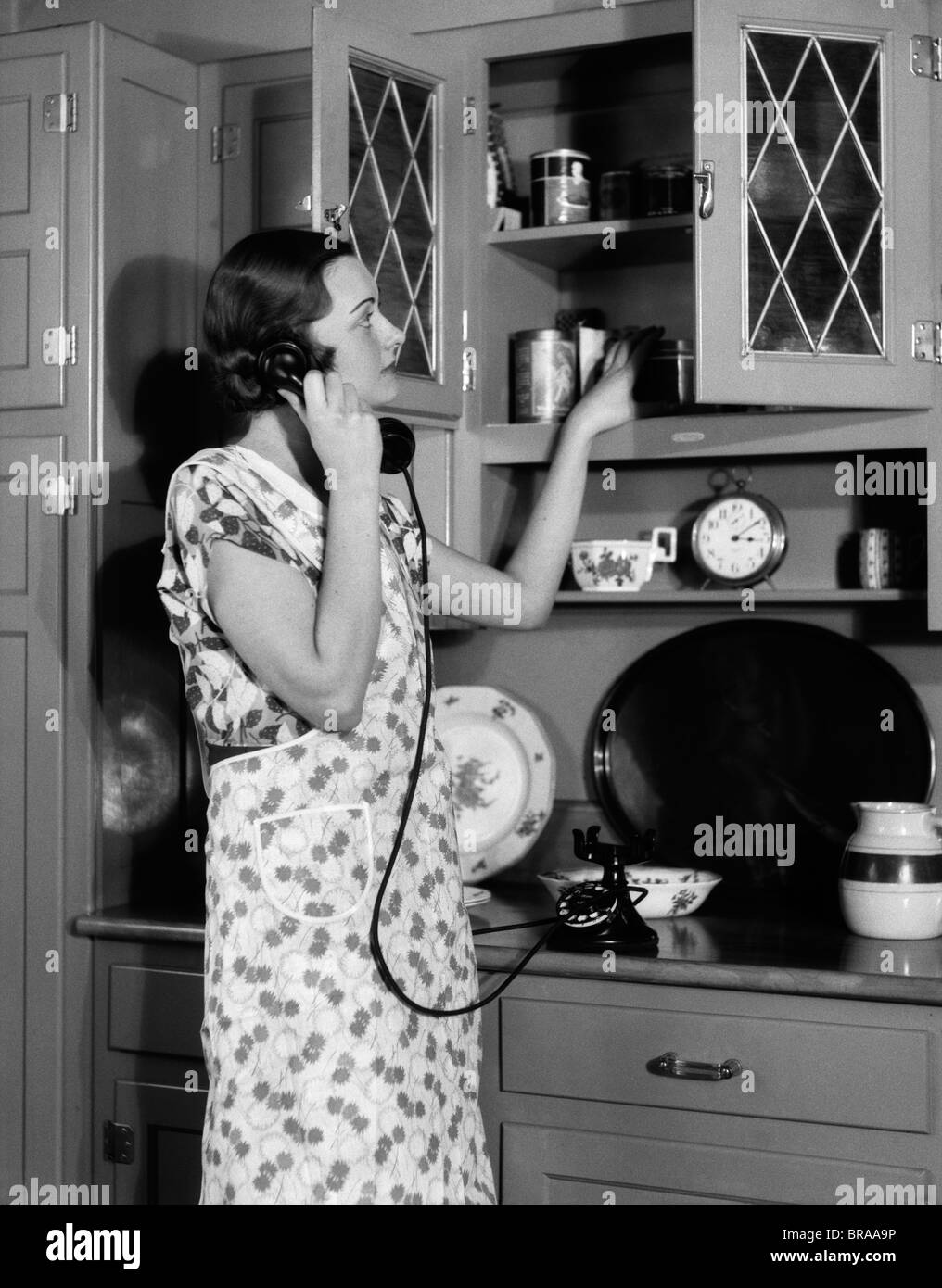 1930s WOMAN HOUSEWIFE WEARING APRON IN KITCHEN TALKING ON TELEPHONE PUTTING FOOD ON CUPBOARD SHELF Stock Photo