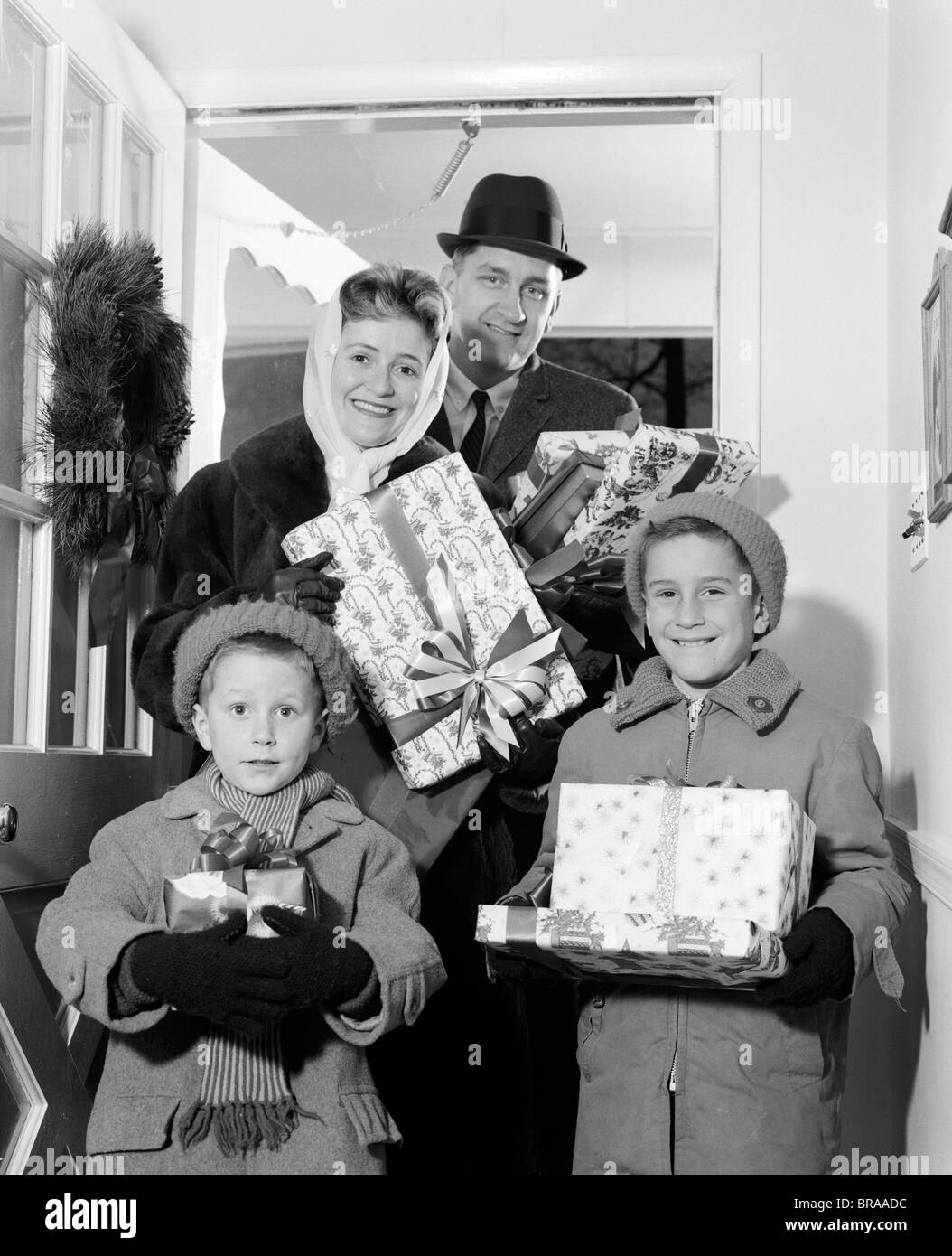 1960s FAMILY STANDING IN DOORWAY OF HOME WEARING WINTER COATS & HATS HOLDING WRAPPED GIFTS Stock Photo