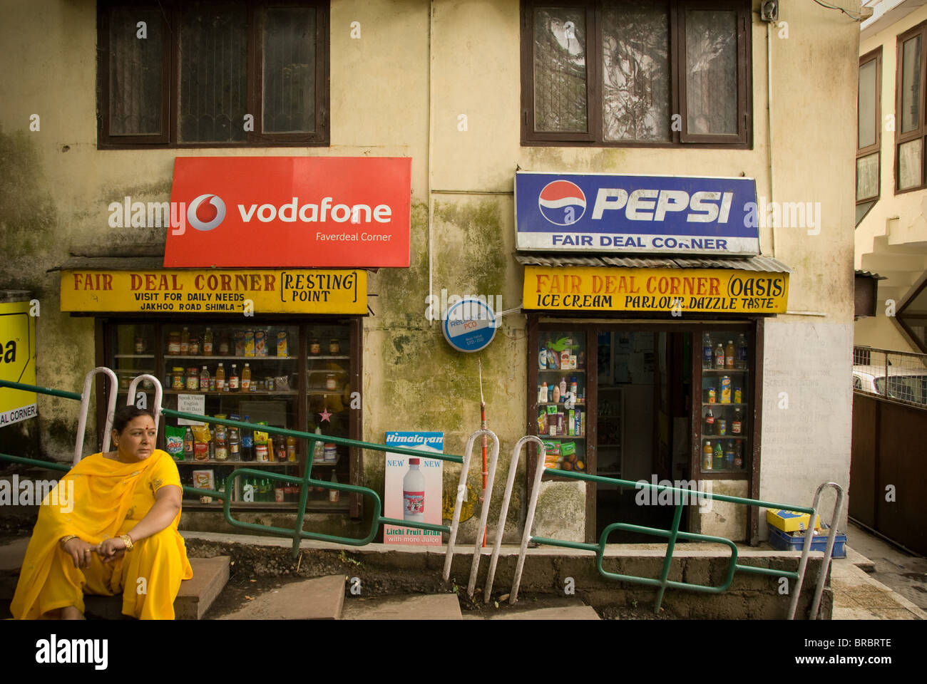 Indian woman in sari sitting down in front of corner shop with Western ad hoardings -Vodafone and Pepsi Stock Photo