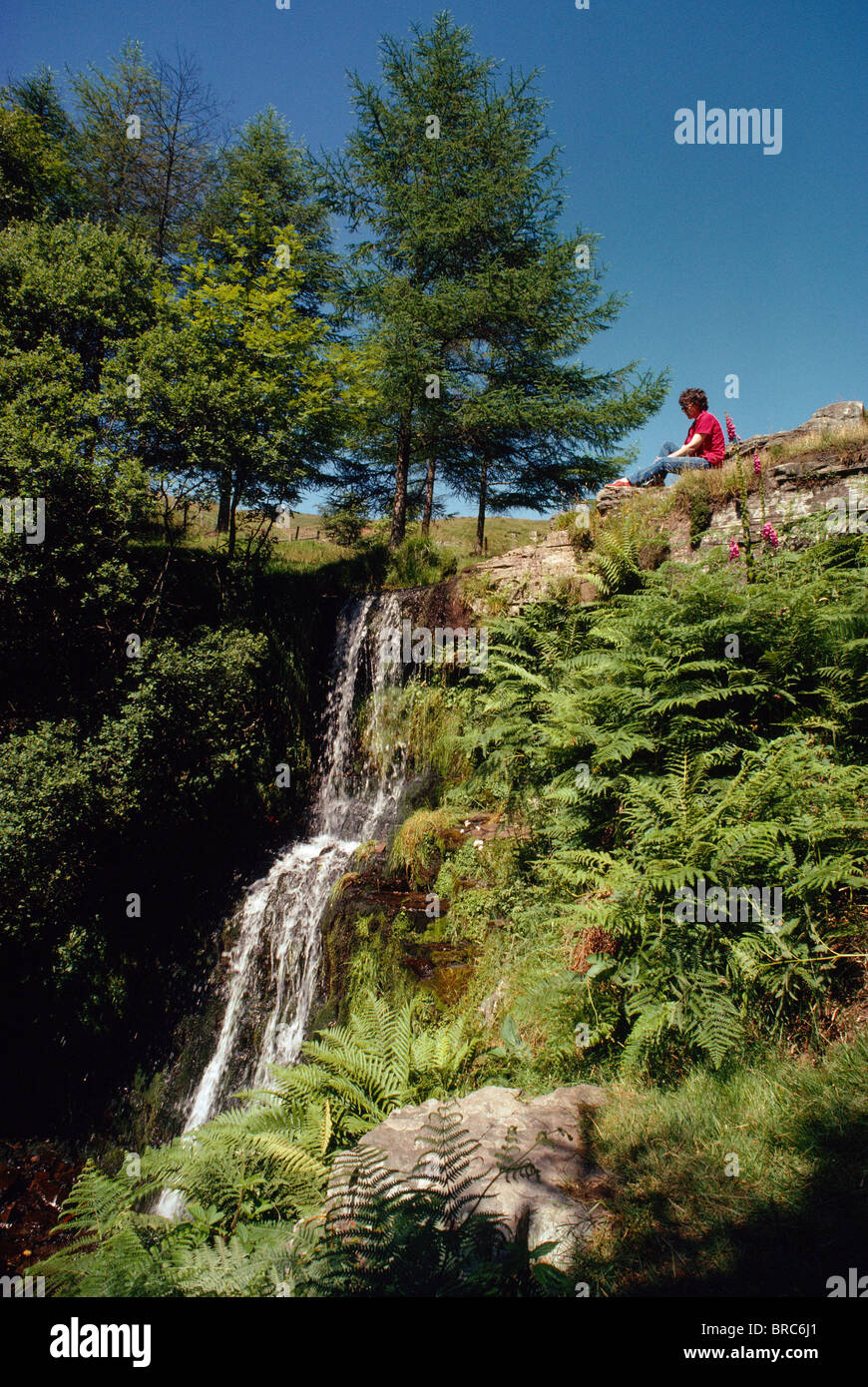 WOMAN IN RED SITTING BY SMALL WATERFALL IN BRECON BEACONS POWYS Stock Photo