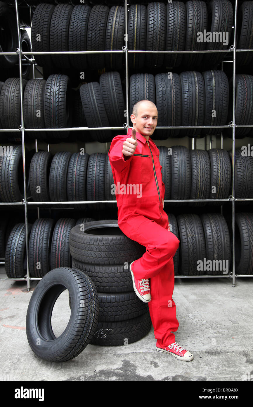 A positive mechanic in a garage standing next to a rack full of tires and making a positive gesture Stock Photo