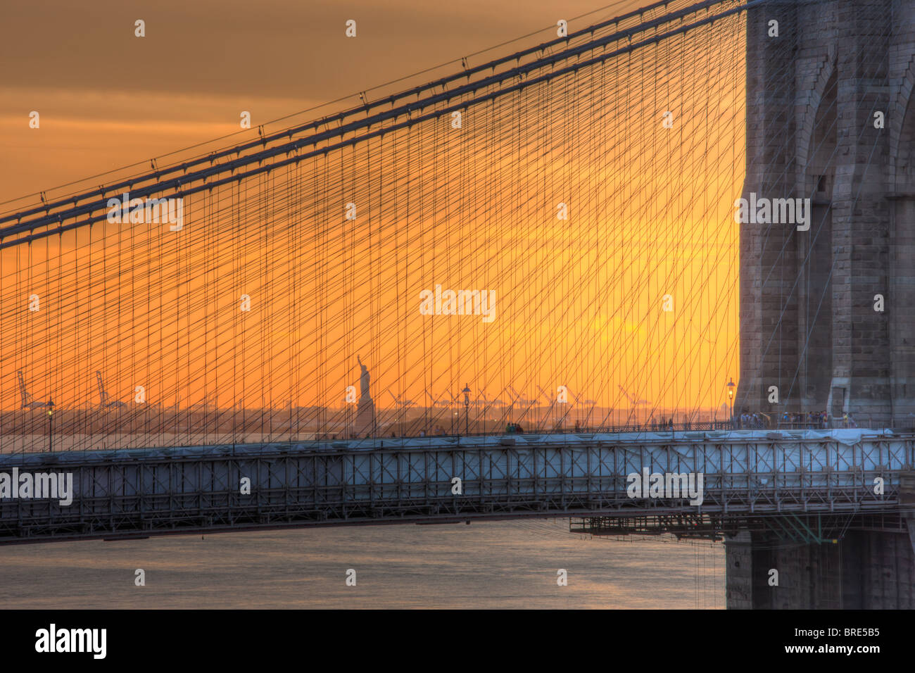 The Brooklyn Bridge with the Statue of Liberty set against the orange sky at sunset. Stock Photo