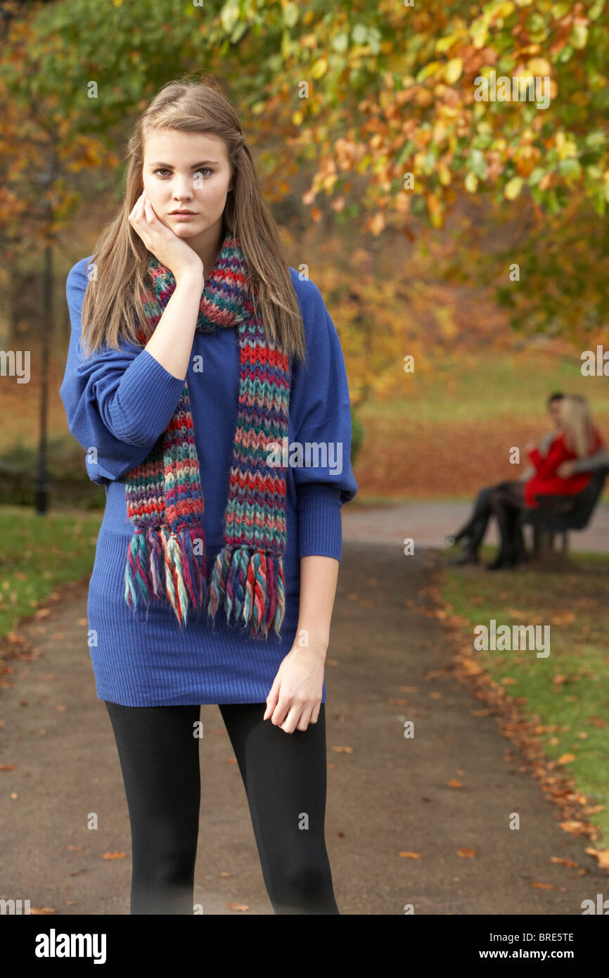 Unhappy Teenage Girl Standing In Autumn Park With Couple On Bench In Background Stock Photo