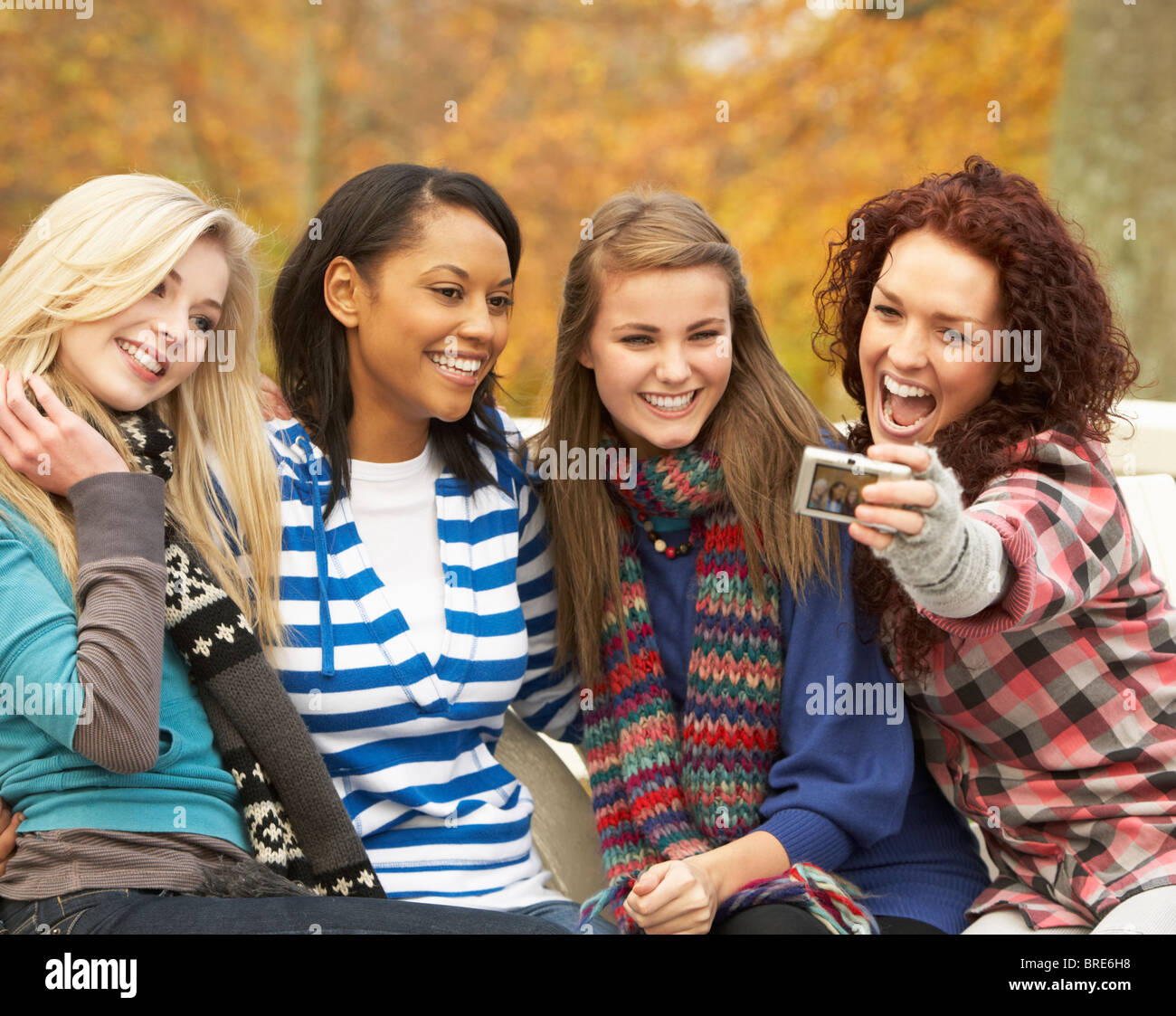 Group Of Four Teenage Girls Taking Picture With Camera Sitting On Bench In Autumn Park Stock Photo