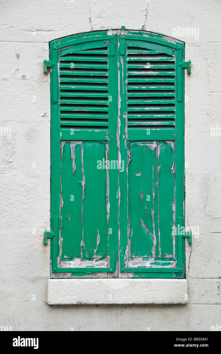 A window with closed green shutters and peeling weathered paint, France Stock Photo