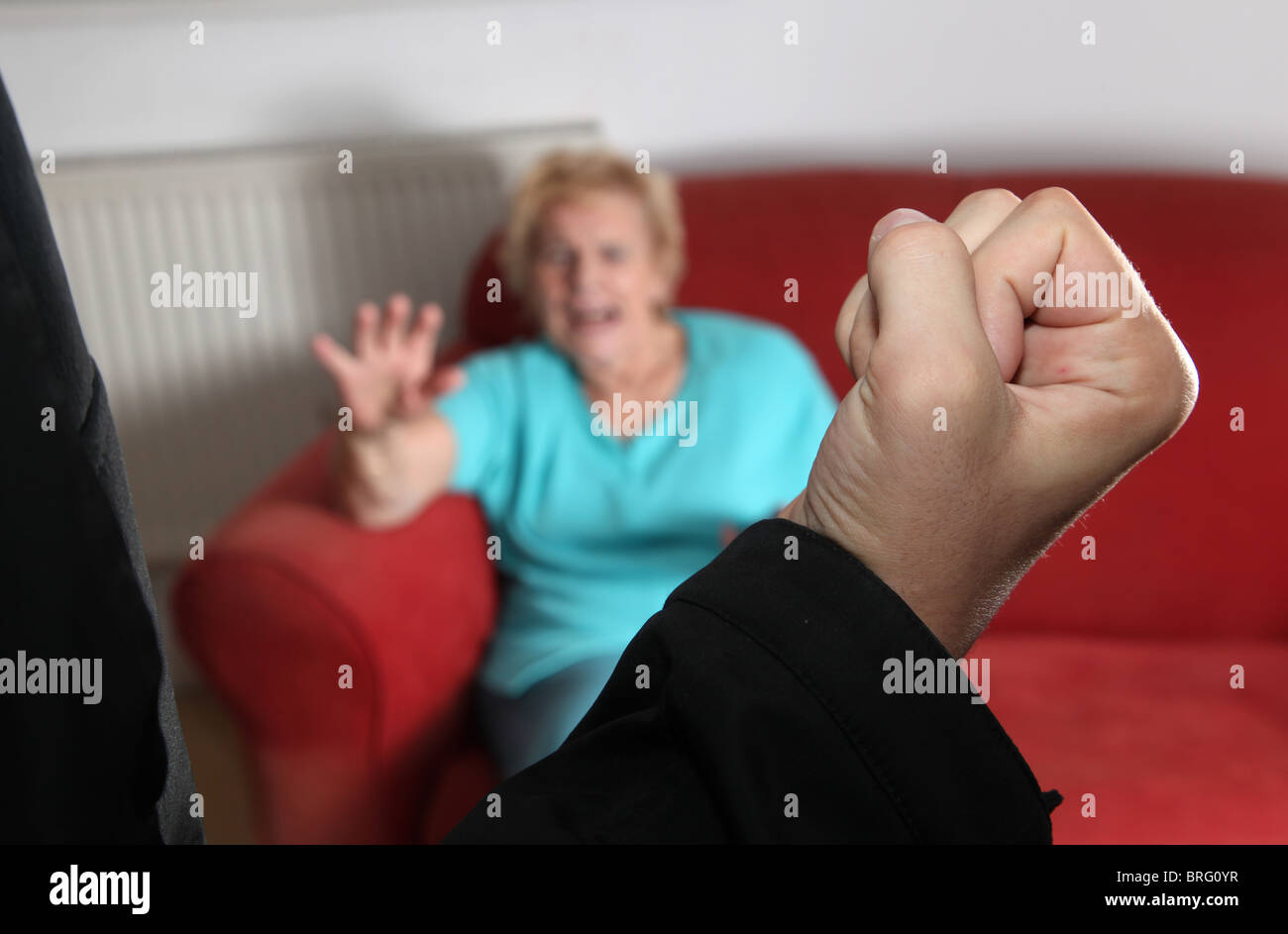 Male fist raised aimed towards an elderly woman sitting on a sofa at home Stock Photo