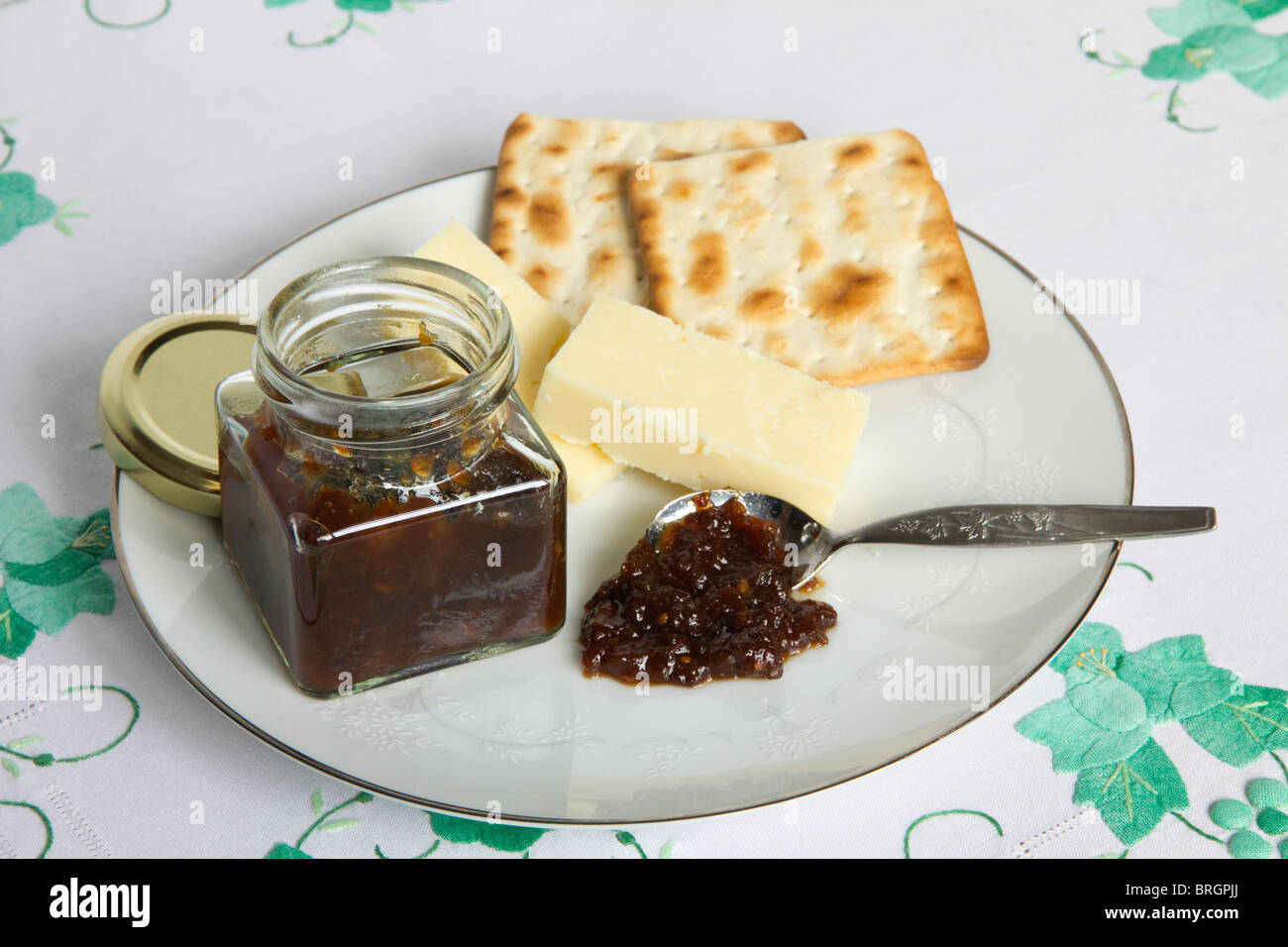A snack of Cheese, biscuits and a jar of chutney on a white plate placed on tablecloth with green applique embroidery Stock Photo