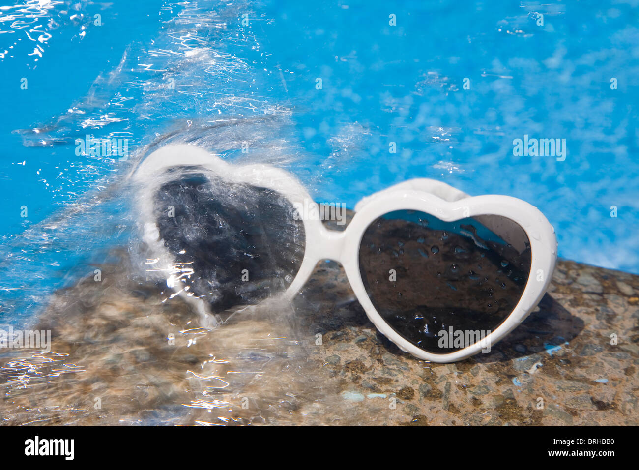 Heart shaped sunglasses by a bright blur swimming pool with splashing water Stock Photo