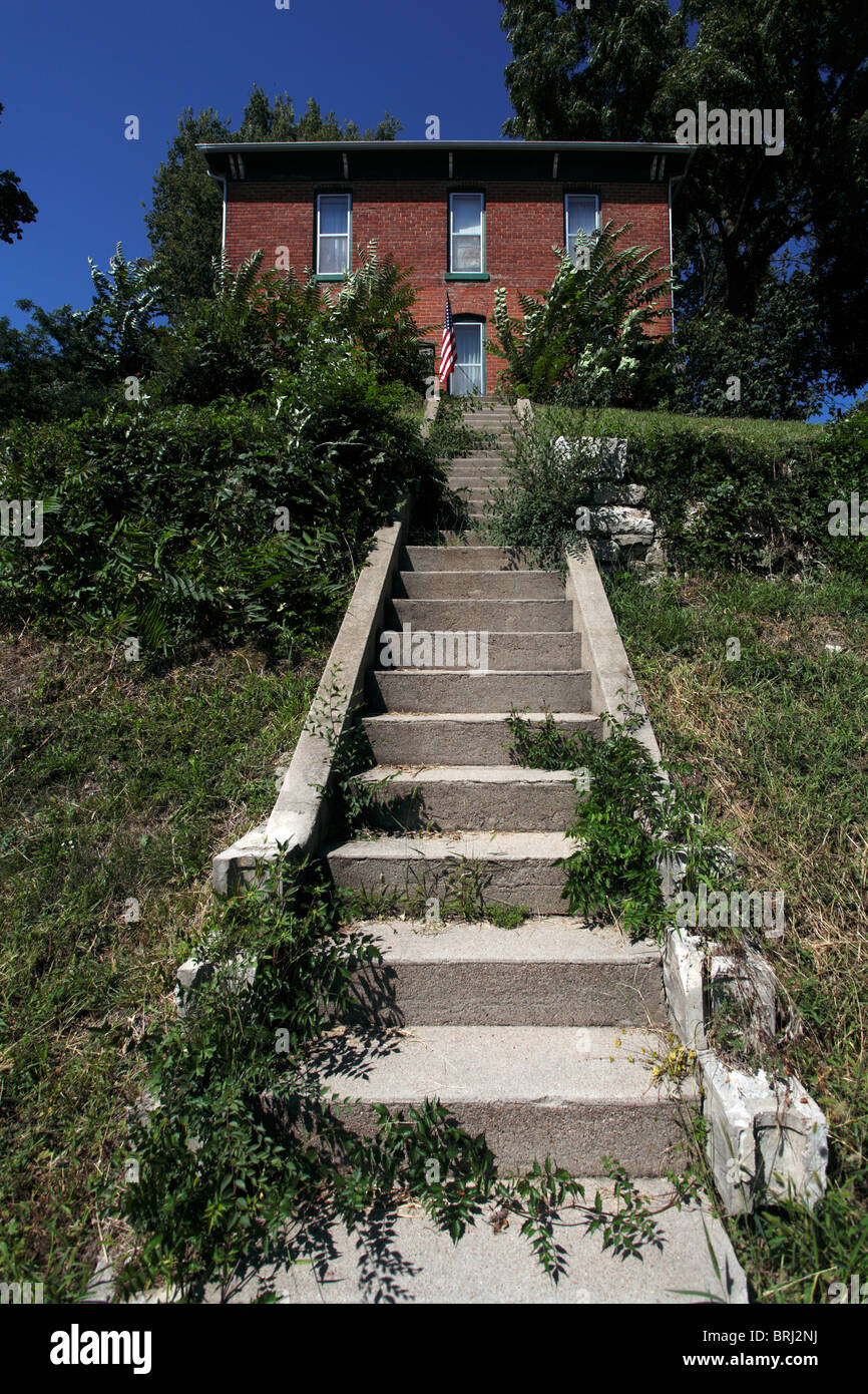 A red brick house on the top of a hill, with a concrete staircase leading up and American flag near door. Stock Photo