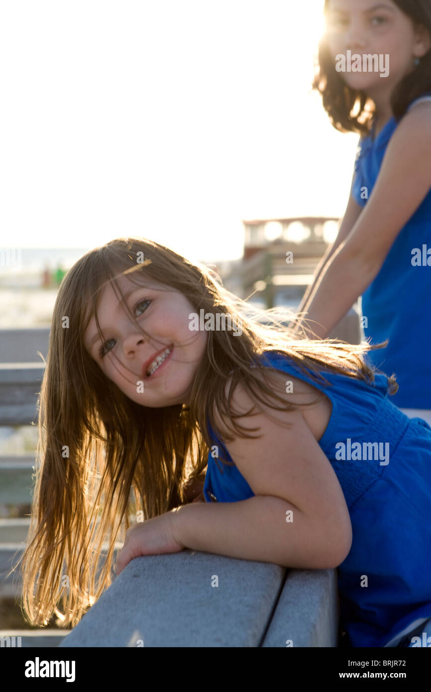 Two little girls on a boardwalk railing are facing the camera with the sun in their hair. Stock Photo