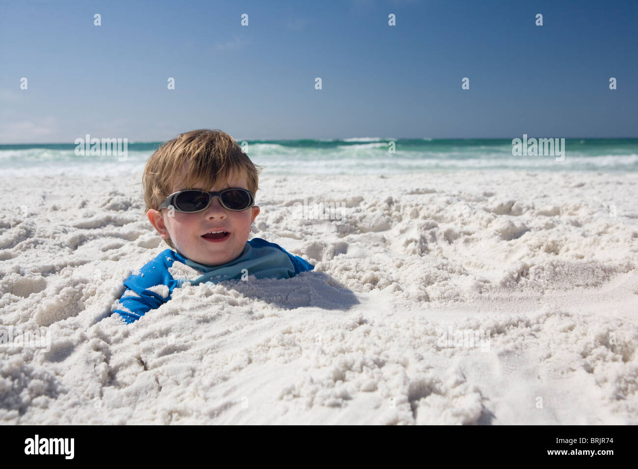 A little boy with sunglasses is buried in the sand up to his head with blue sky and ocean in the background. Stock Photo