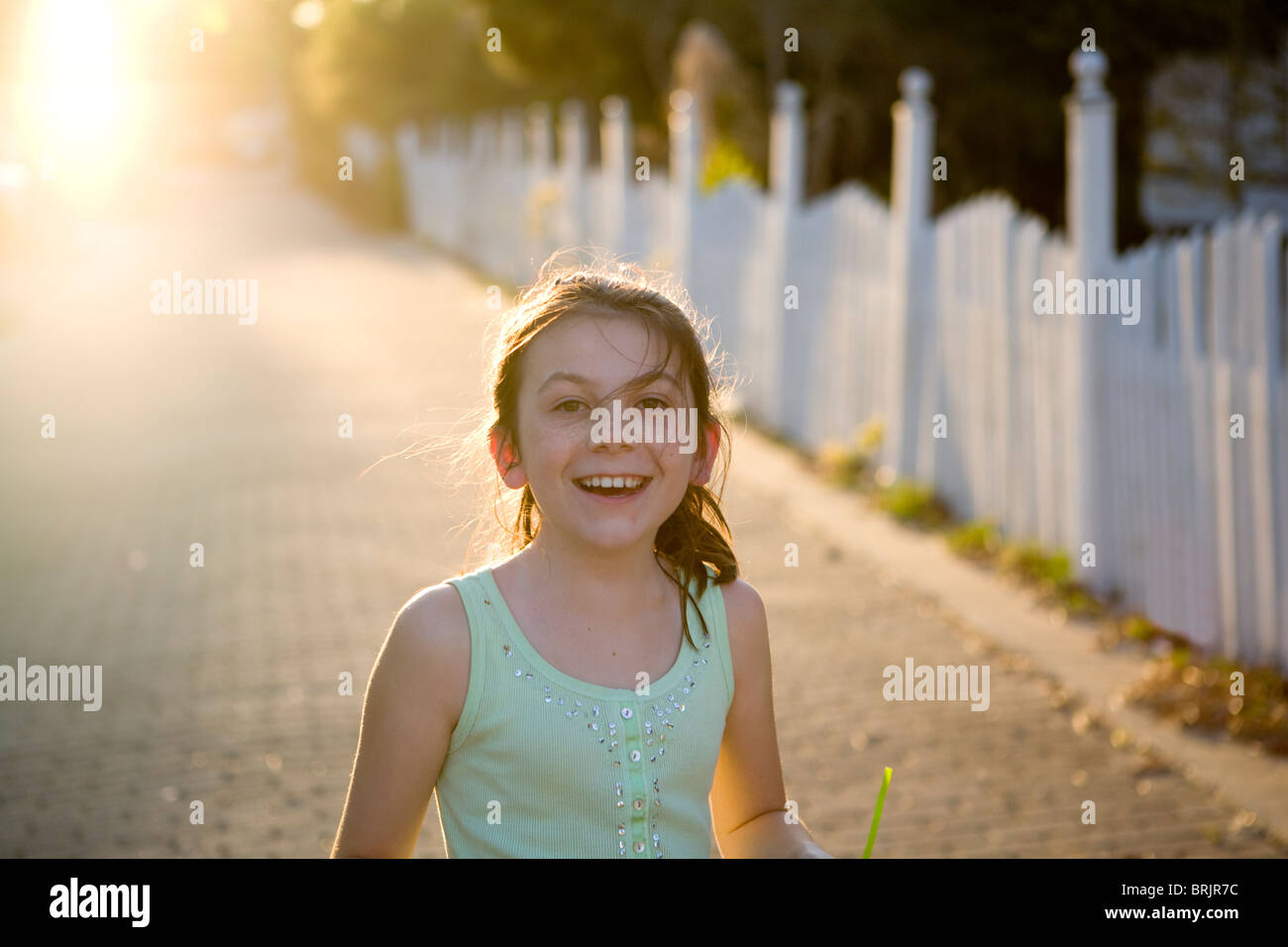 A freckled girl is smiling with a picket fence and sun in the background. Stock Photo