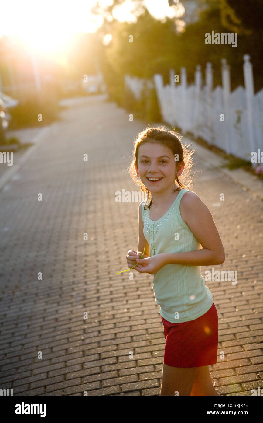 A little girl wearing shorts is smiling at the camera with a picket fence and sun in the background. Stock Photo
