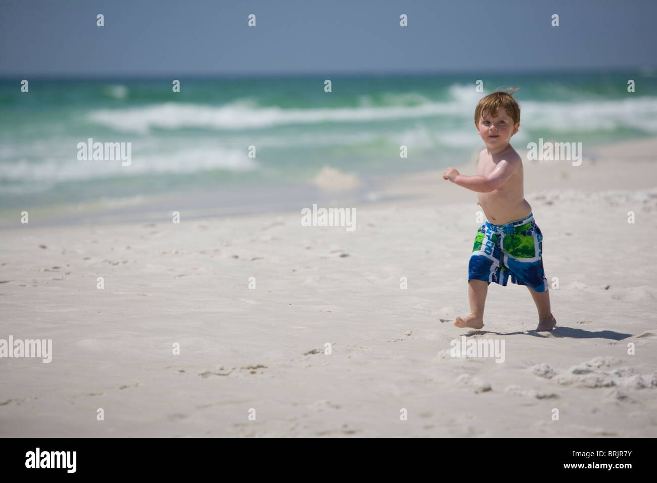 A little boy struts down the beach with the ocean in background. Stock Photo