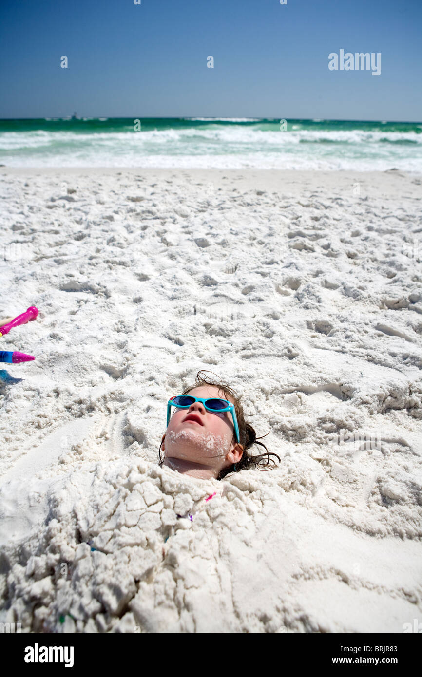 A little girl is buried in the sand up to her head at the beach with the ocean in the background. Stock Photo