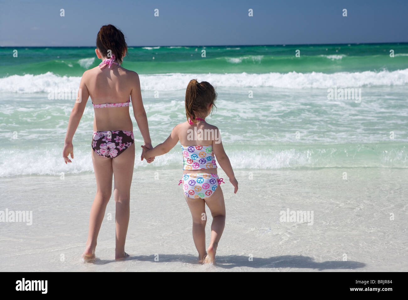 Two little girls in bikinis are holding hands at shoreline with the ocean in the background. Stock Photo