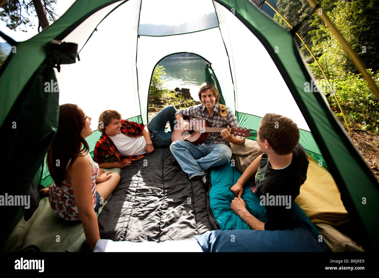 Young adults camping listen to a friend play guitar inside the tent next to a lake in Idaho. Stock Photo