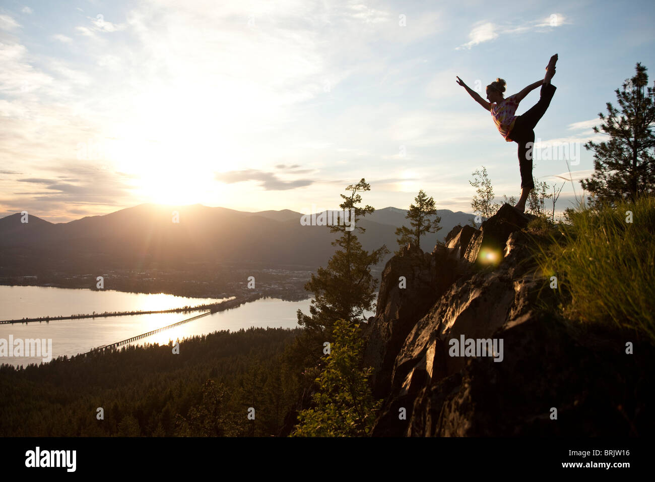 Young woman doing yoga at the top of a mountain overlooking a small town and large lake. Stock Photo