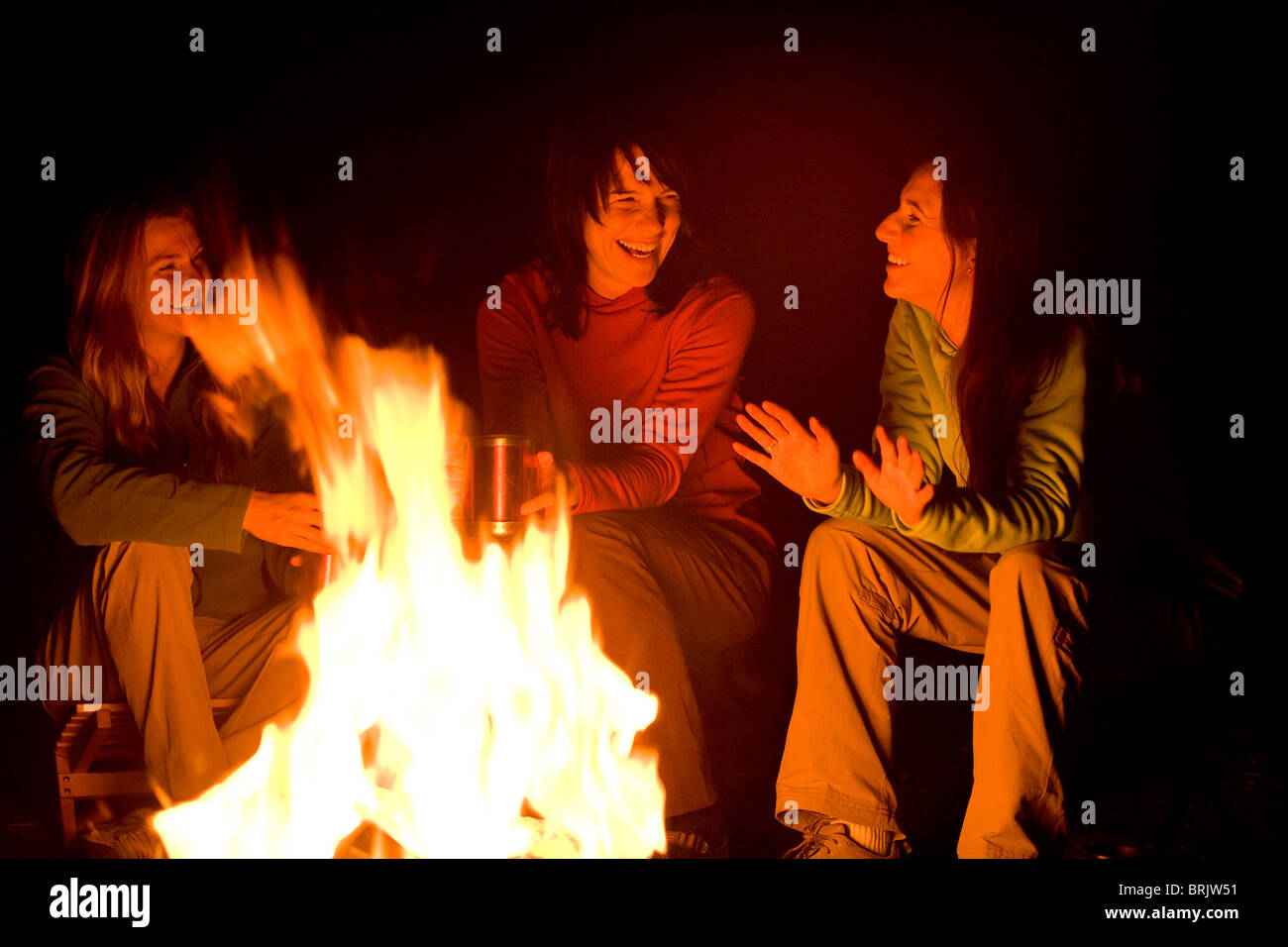 Three women talk and laugh together around a campfire. Stock Photo