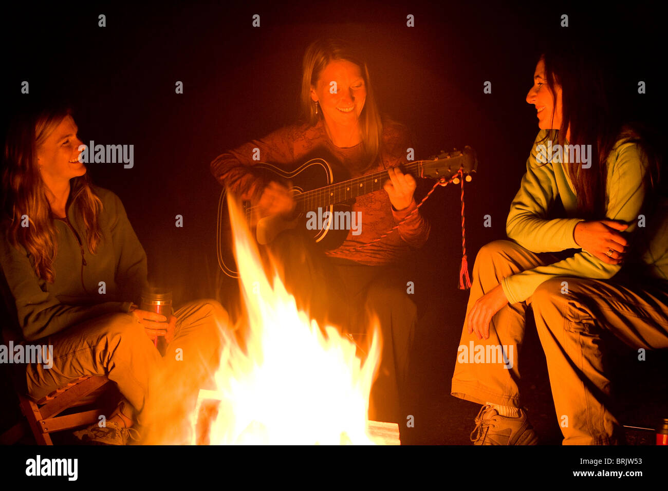 Two women listen, as a third plays the guitar beside a campfire. Stock Photo
