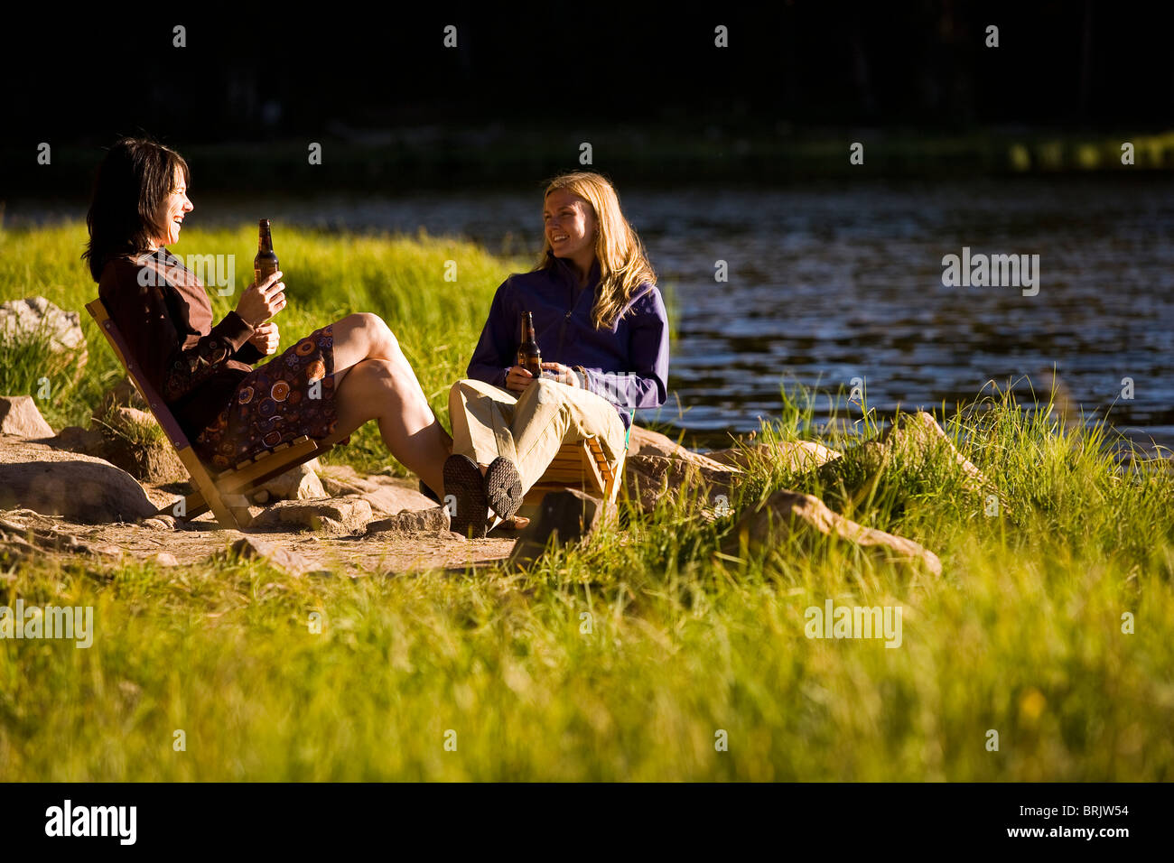 Two women enjoy one another's company and a beer on the shore of a mountain lake. Stock Photo