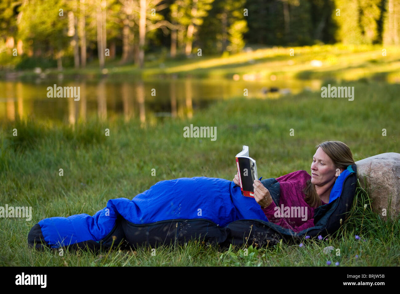 A woman reads in a sleeping bag beside a mountain lake. Stock Photo