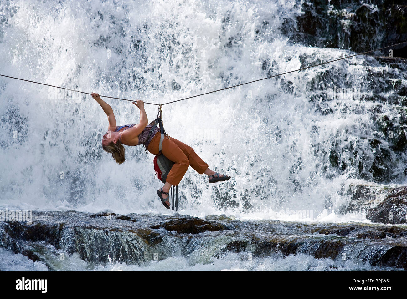 A woman crosses a waterfall using a Tyrolean Traverse on a rope. Stock Photo