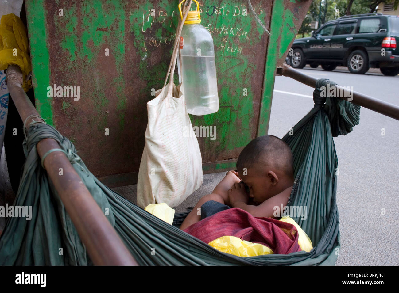 A young boy is sleeping in a hammock tied to a Cintri garbage collection cart on Norodom Boulevard in Phnom Penh, Cambodia. Stock Photo