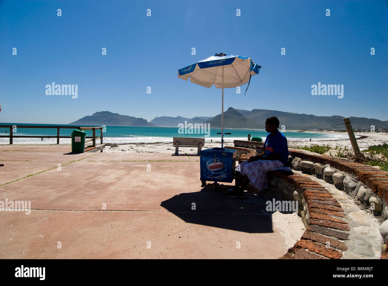 Ice Cream seller sitting in the shade of an umbrella in the parking area at Kommetjie beachfront Stock Photo