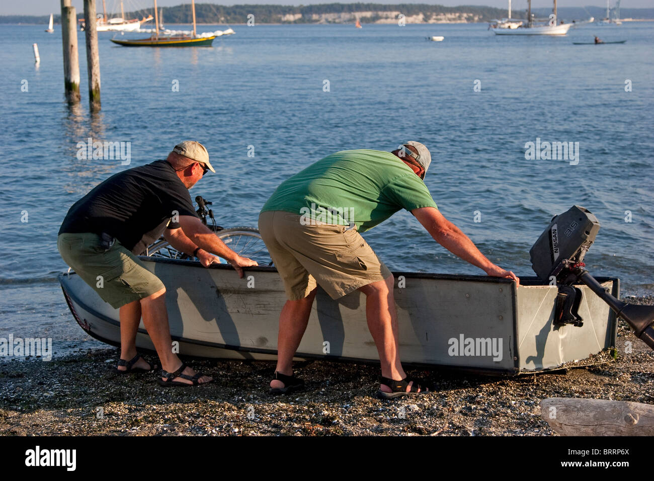Two men heave and pull a small wooden dingy boat from the beach into the water, Port Townsend, Washington, USA Stock Photo