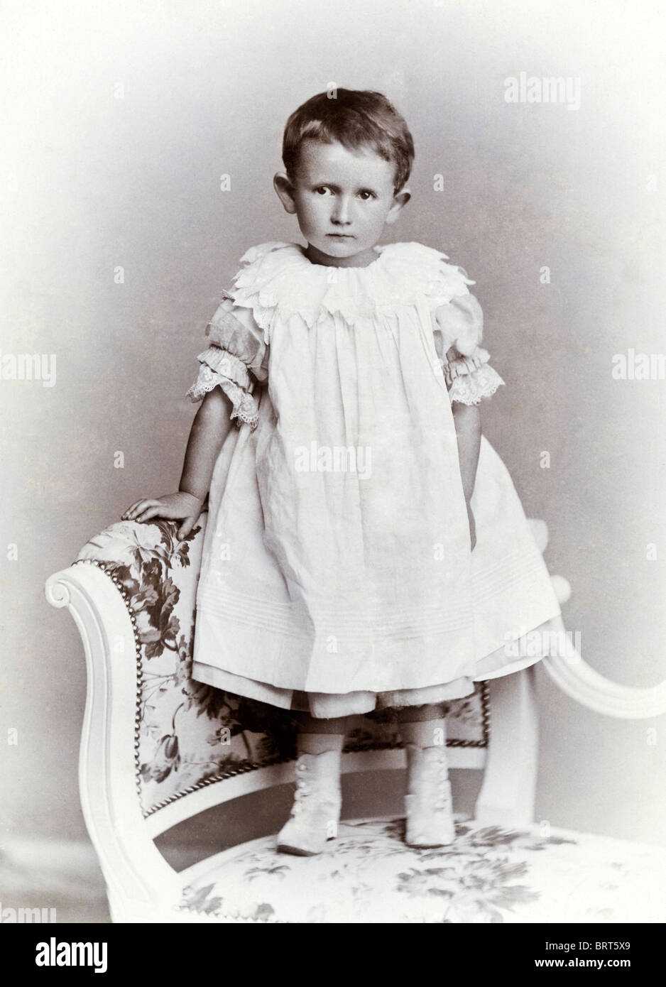 Studio portrait of Victorian child with short hair wearing a smock dress standing on chair circa 1885 Stock Photo
