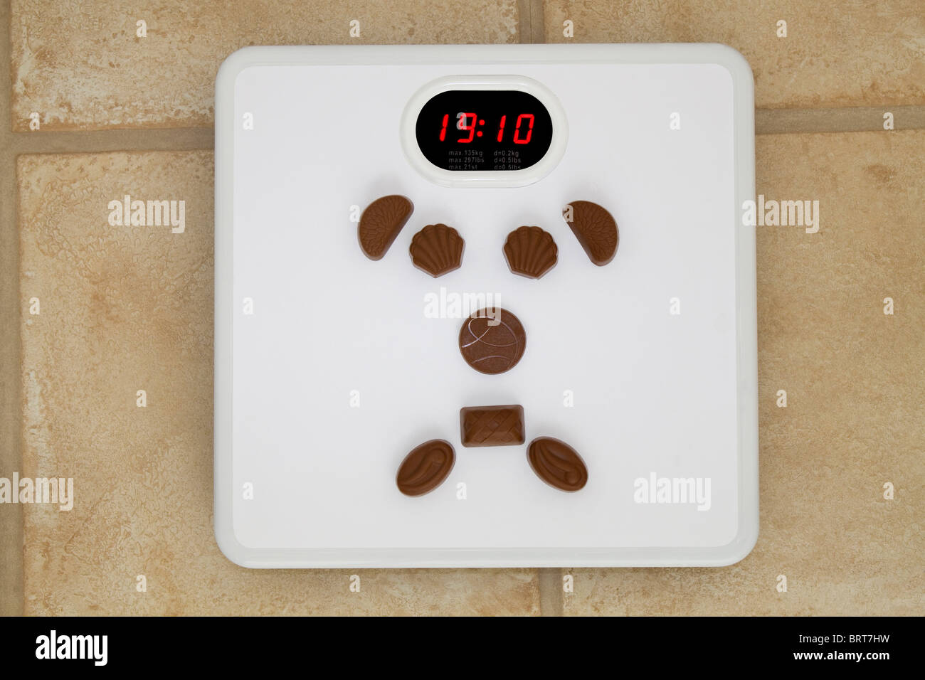 A set of bathroom scales which has chocolates in the shape of a sad face reads, 19 stone 10 pounds Stock Photo