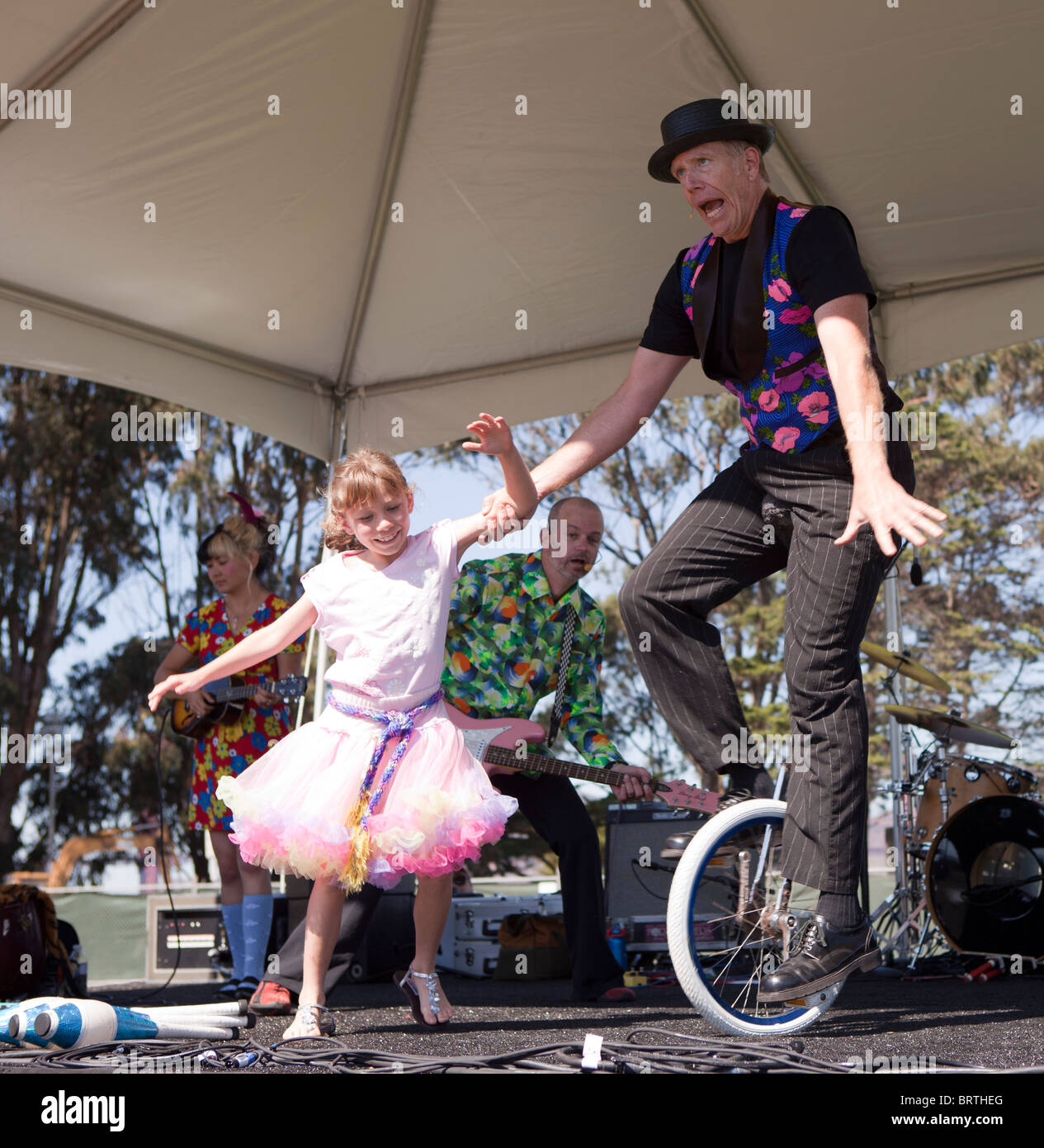 A man rides a unicycle with the help of a little girl at an outdoor music festival Stock Photo