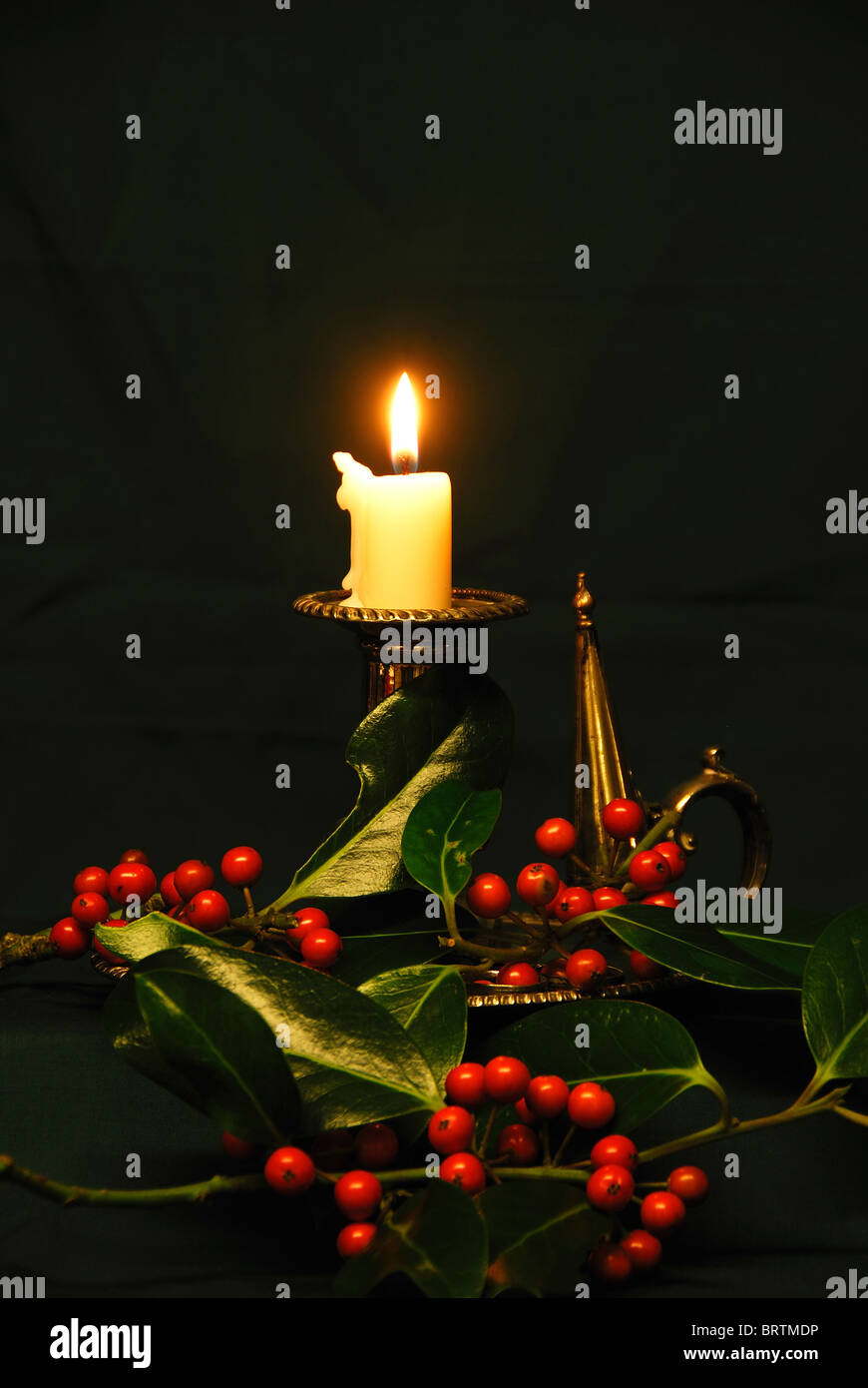 A silver candle holder with a burning candle and red holly berries. A ...