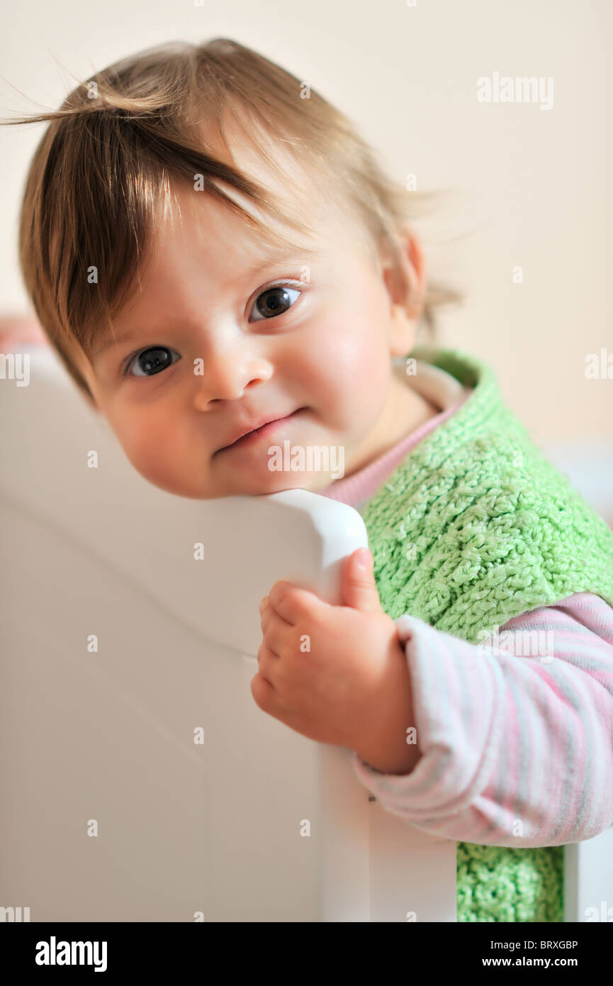 A toddler girl leans her head on the headboard of her white cot