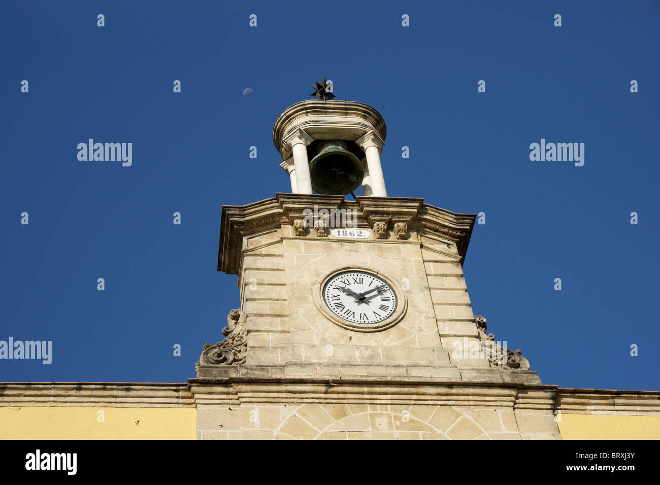 Old Bell tower with clock in Jerez. Daytime, though the Moon is visible in the clear blue sky. Stock Photo