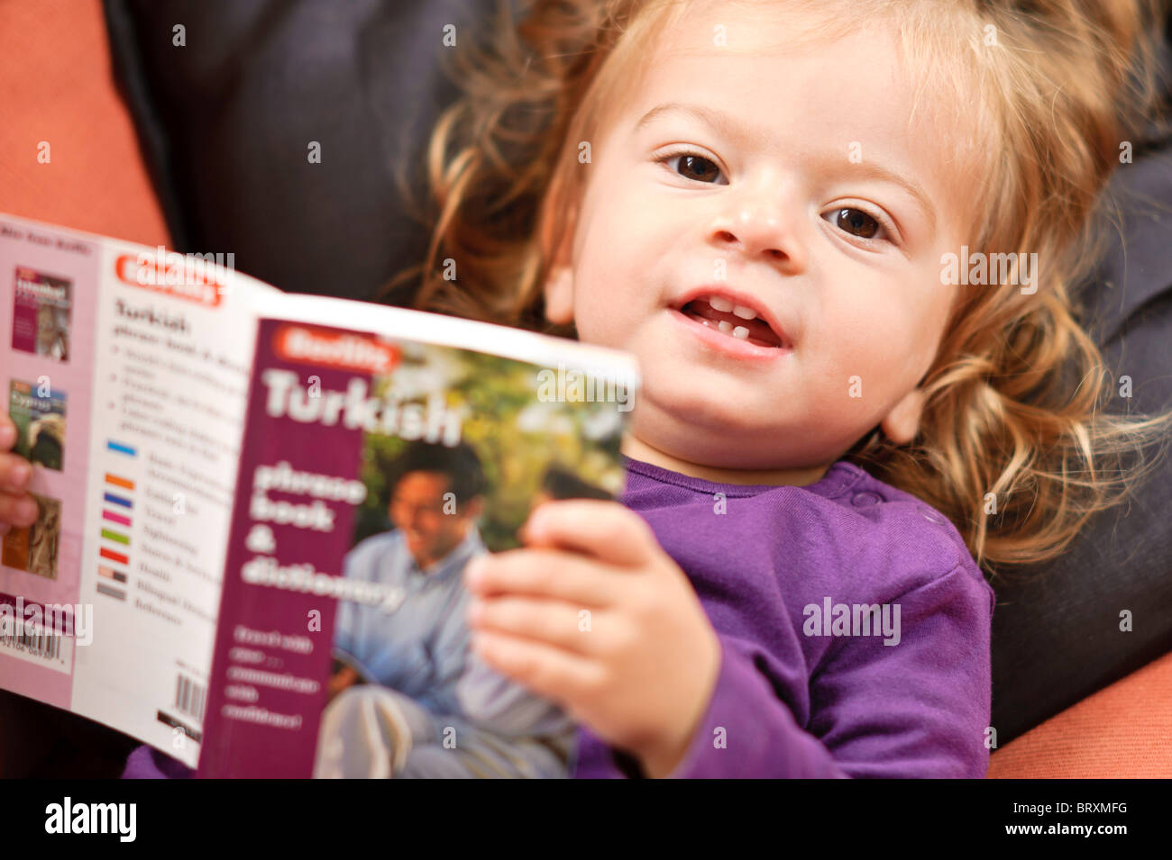 A toddler girl relaxing on a sofa reads a Turkish language study book. Stock Photo
