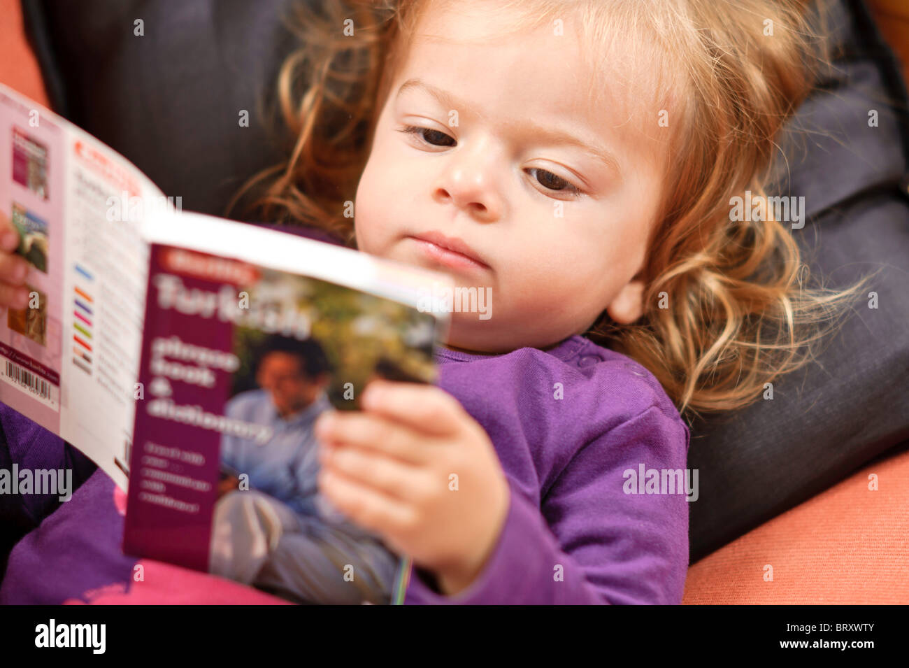 A toddler girl relaxing on a sofa reads a Turkish language study book. Stock Photo