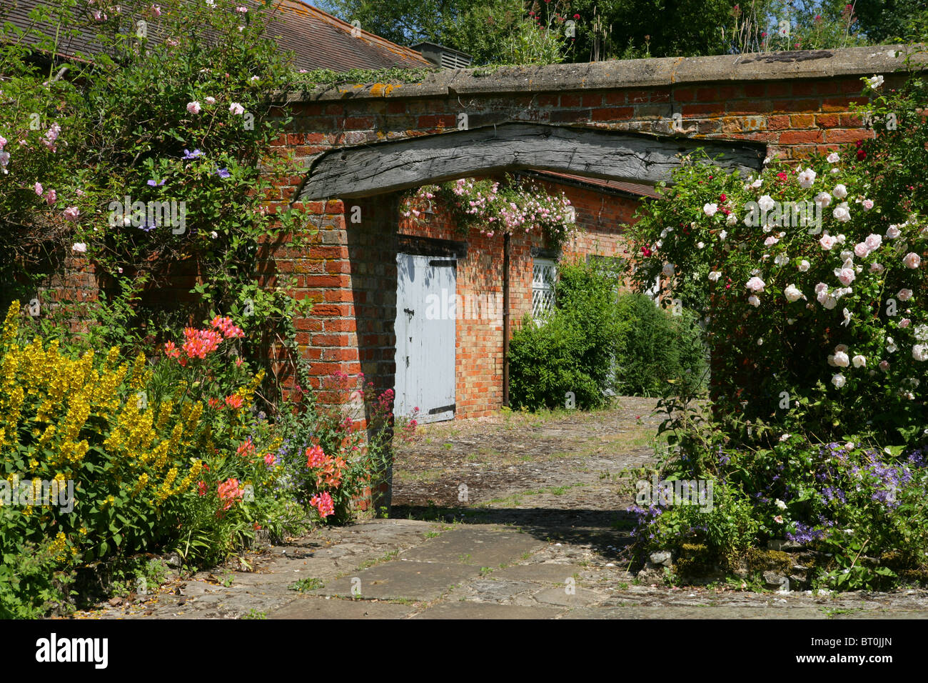 Old farmhouse brick and wooden archway leading into stable yard with summer roses and hanging baskets, England. Stock Photo