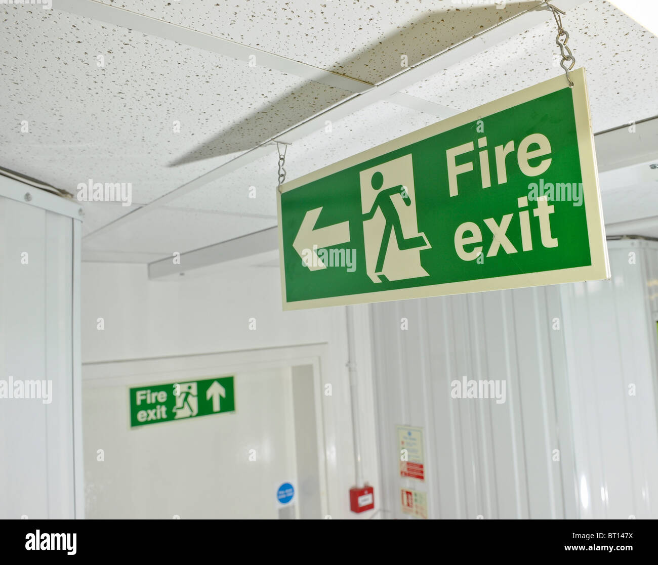 Green Fire Exit Sign Hanging From Ceiling Pointing To Escape Exit