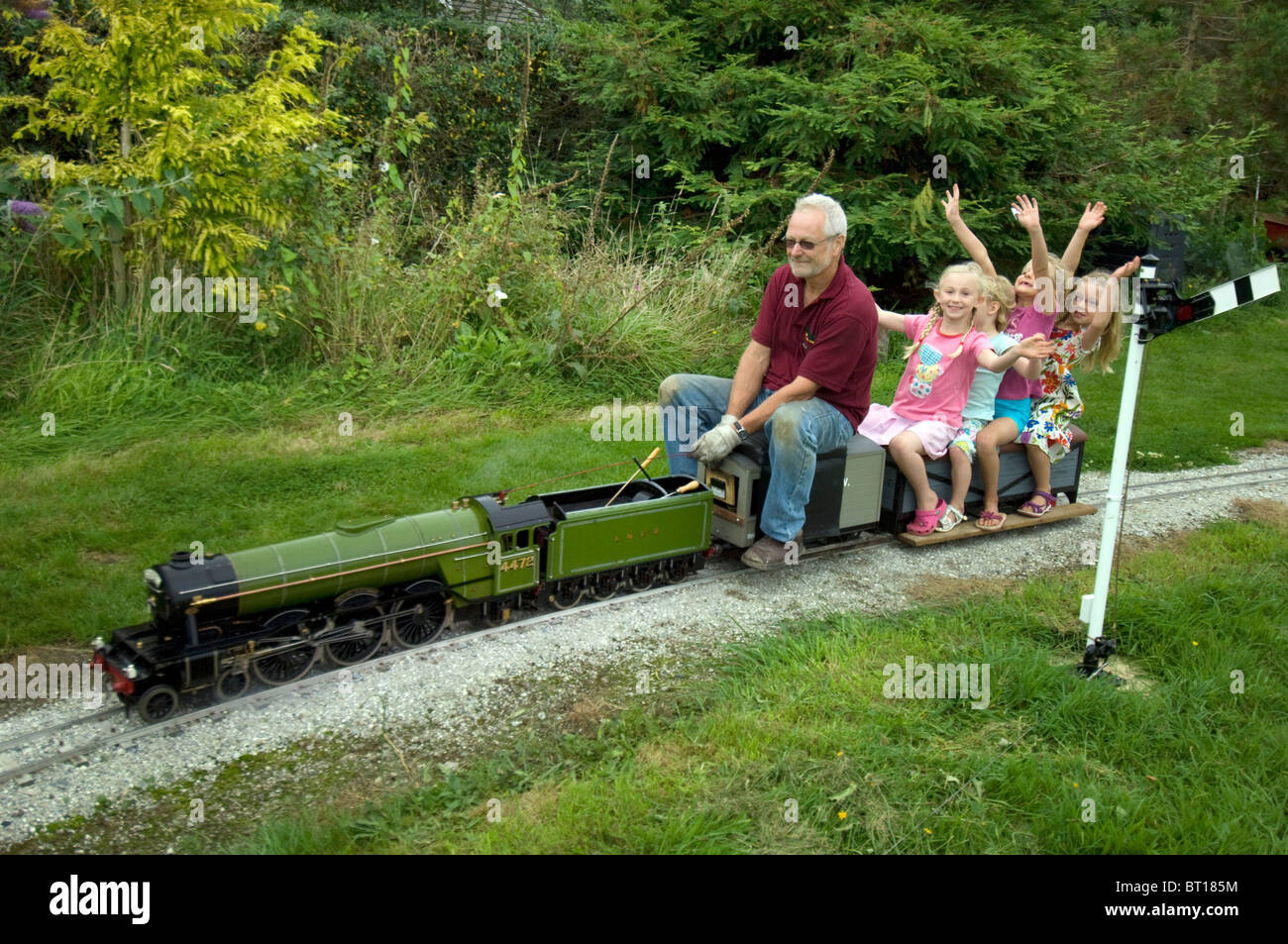 Three little girls waving as they ride a 5 inch gauge model  of 'Flying Scotsman' LNER train driven by a man with grey hair. Stock Photo